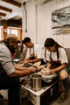 Three individuals, two women and a man, wearing aprons and collaborating on pottery projects at a pottery wheel in a rustic workshop.