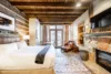 A cozy, rustic bedroom featuring a large bed with white bedding, wooden beam ceiling, stone fireplace, and a patterned rug. a seating area with a chair by the window and modern amenities add comfort.