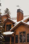 A wooden cabin covered in snow with a prominent chimney, nested in a wintry landscape with cloudy skies.