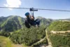 A person in a harness and helmet ziplines over a lush green mountain landscape under a clear blue sky.