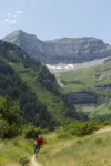 A cyclist in a red shirt rides along a trail through a lush green valley with towering, layered mountains and patches of snow in the background under a clear sky.