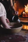 A person pours water from a metal pitcher into a wooden bowl near a cozy, glowing fireplace, with warm ambient lighting enhancing the rustic and peaceful setting.