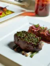 A gourmet plated meal featuring a steak topped with green herbs, served over grilled asparagus, with a square plate of chicken and vegetables in the background.