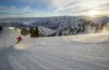 A skier in a pink jacket descends a pristine snowy slope at sunrise, with a panoramic view of snow-covered mountains under a clear sky in the background.