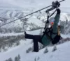 A person zip-lining over a snowy landscape with mountains in the background, raising one arm in excitement. they are dressed in winter gear, including a helmet and goggles.