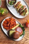 A top-down view of a meal with a burger on a sesame seed bun, sweet potato fries, and fish tacos on a wooden table, accompanied by a mojito cocktail.