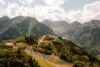 Lush green mountains under a clear sky, with a dirt road winding through the hills and a small building perched on a distant hilltop.