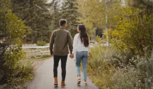 A couple walking hand-in-hand along a park path surrounded by lush greenery and trees, viewed from behind.