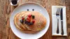 A stack of pancakes topped with fresh strawberries, blueberries, and raspberries, dusted with powdered sugar, served on a wooden table with maple syrup on the side and a white napkin with silverware.