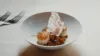 A gourmet dish featuring scallops and grains served in a white bowl, garnished with herbs and delicate, crisp wafer, presented on a white tablecloth with silverware on the side.