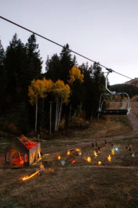A ski lift chair ascends over a forest with colorful autumn leaves. below, an illuminated halloween decoration display includes lit pumpkins and a small haunted house structure at twilight.