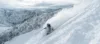 A skier descends a steep, snowy slope with a cloud of powder trailing behind, surrounded by rugged, snow-covered mountains under a cloudy sky.