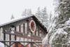 A cozy, snow-covered cabin with a festively decorated wreath on the facade, surrounded by snowy pine trees, creating a picturesque winter scene.
