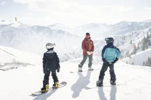 Two young children learning how to snowboard are listening to their instructor.
