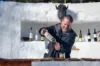 A bearded man pours a bottle of liquor at an outdoor bar made of ice, surrounded by bottles, with a snowy landscape in the background.