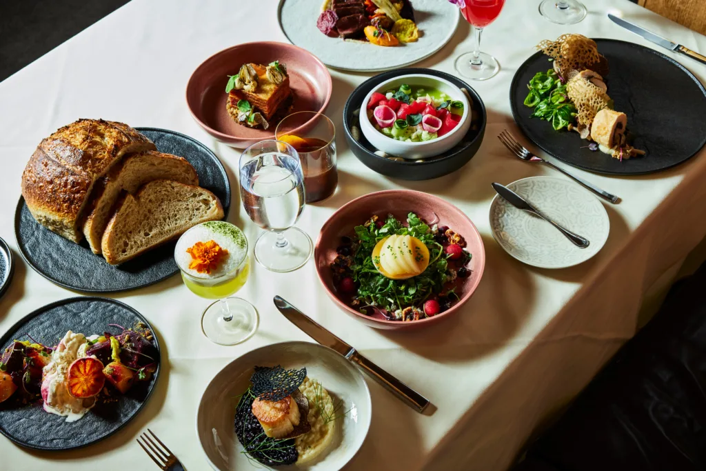 Elegant dining table set with various gourmet dishes including fish, salads, bread, and a poached egg, beautifully presented on colorful plates.