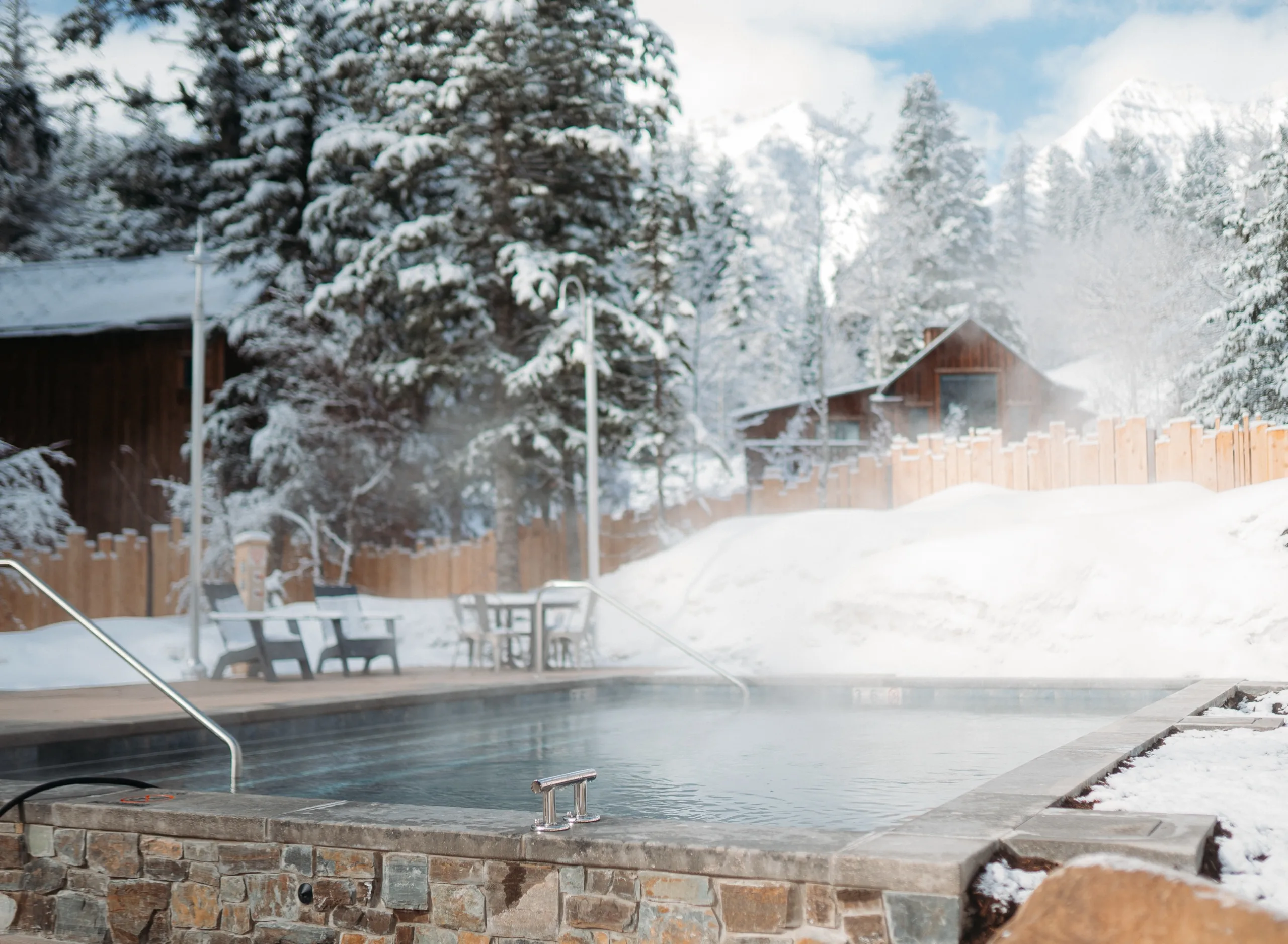 An outdoor heated pool in a snowy landscape with pine trees, steam rising from the water, and a few buildings in the background, under a clear blue sky near some of the best Sundance restaurants.