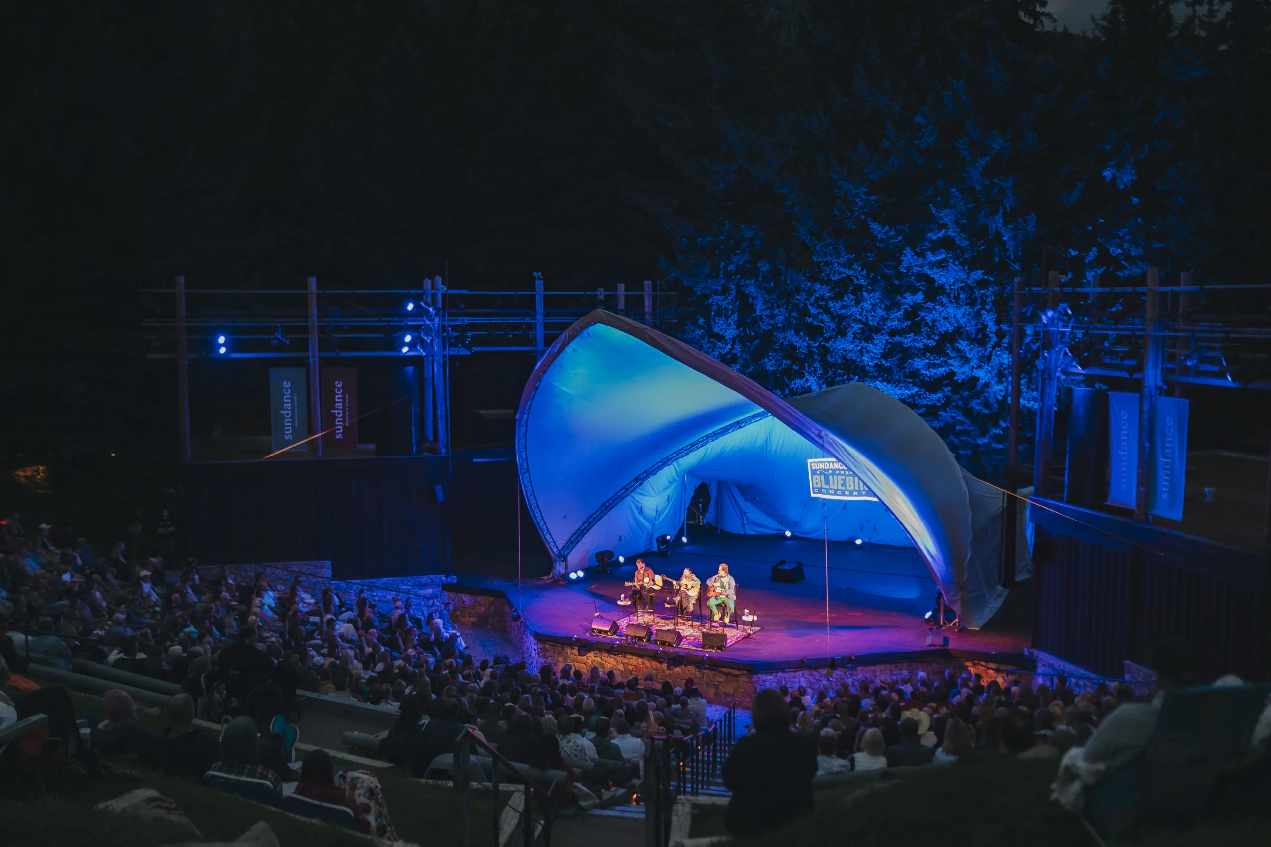 An audience watches a nighttime outdoor concert at an amphitheater. The stage, illuminated with blue lighting, features a group of musicians performing under a white canopy. Trees and dark night sky form the backdrop, creating a serene, open-air setting.