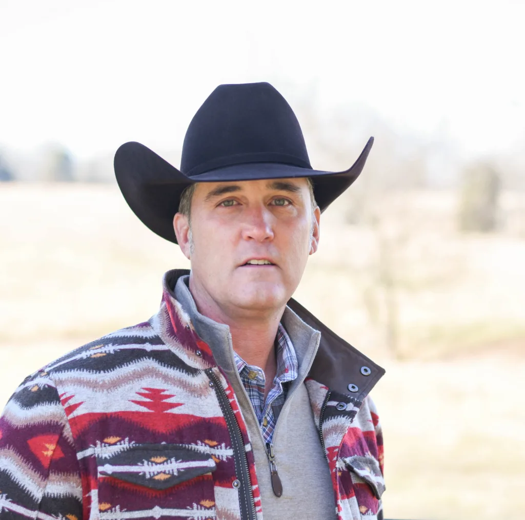 A man wearing a cowboy hat, patterned jacket over a shirt, stands outdoors with a field in the background. his expression is serious.