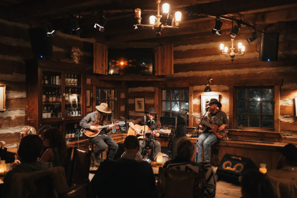 A cozy, warmly lit log cabin hosts a live musical performance. Three musicians play guitars and sing, seated in front of a small audience. The rustic interior features wooden beams, a chandelier, and shelves stocked with bottles, creating an intimate atmosphere.