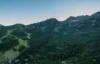 A scenic view of a lush green mountainous landscape with dense forests and scattered houses in a valley. The mountain peaks rise in the background under a clear, blue sky.