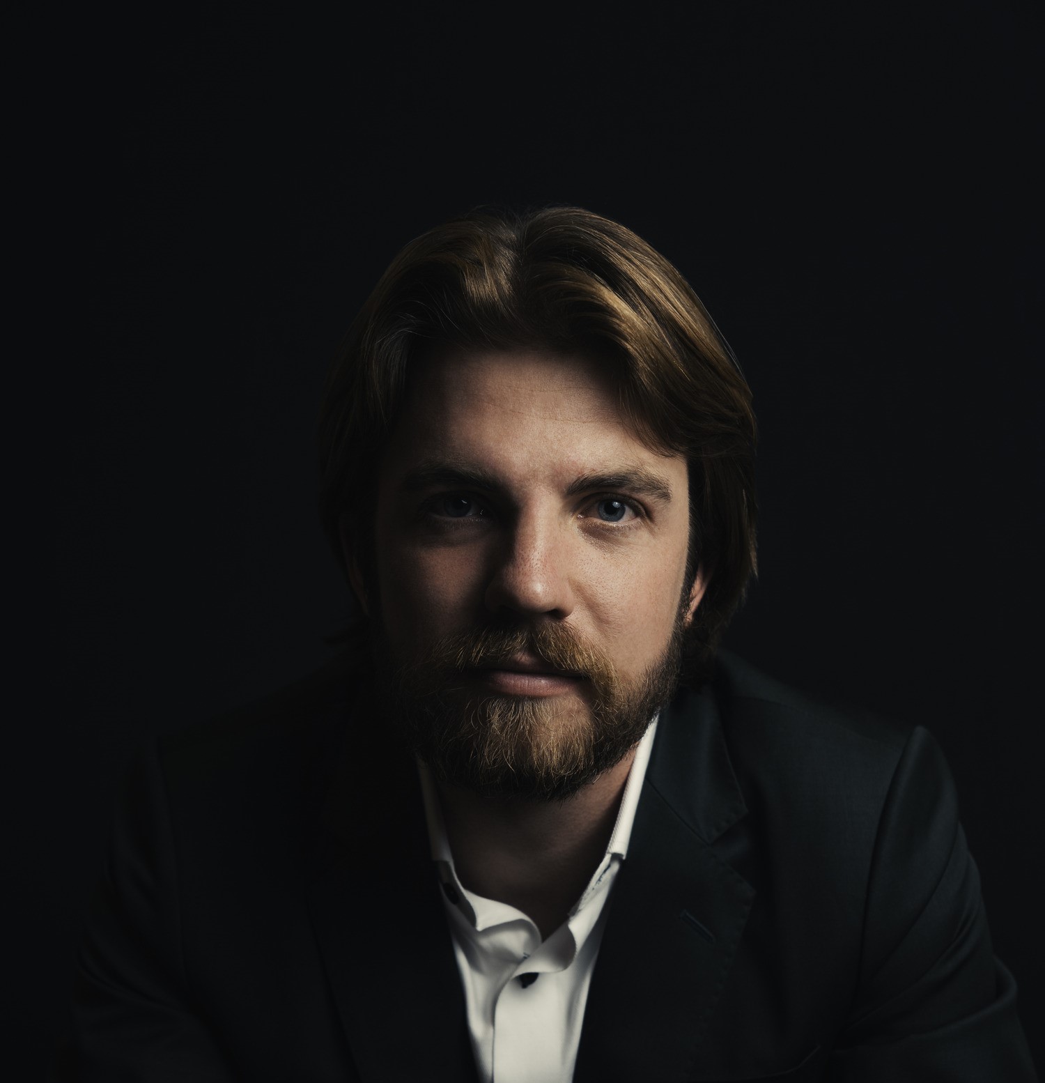 A young man with medium-length hair, a beard, and mustache, wearing a dark suit jacket over a white shirt, sits against a dark background. He gazes directly at the camera with a neutral expression, and the lighting highlights his face, casting subtle shadows.