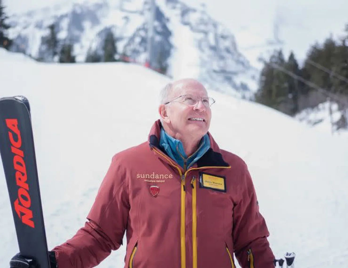 An older man in a red jacket with a "Nordica" ski in his right hand stands against a snowy mountain backdrop. He wears glasses and smiles, looking upwards. The jacket has "Sundance" and other patches on it. Pine trees and snow-covered slopes are visible behind him.
