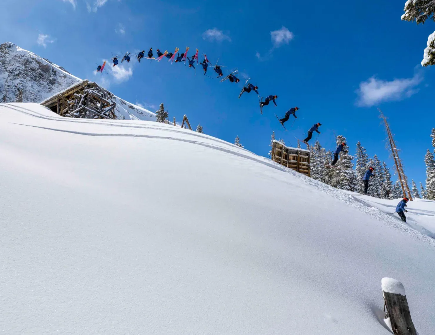 A snowboarder performs a high jump off a wooden ramp in a snowy landscape, captured in multiple frames showing their aerial progression. The scene is set against a backdrop of clear blue sky, snow-covered trees, and a steep snowy slope.