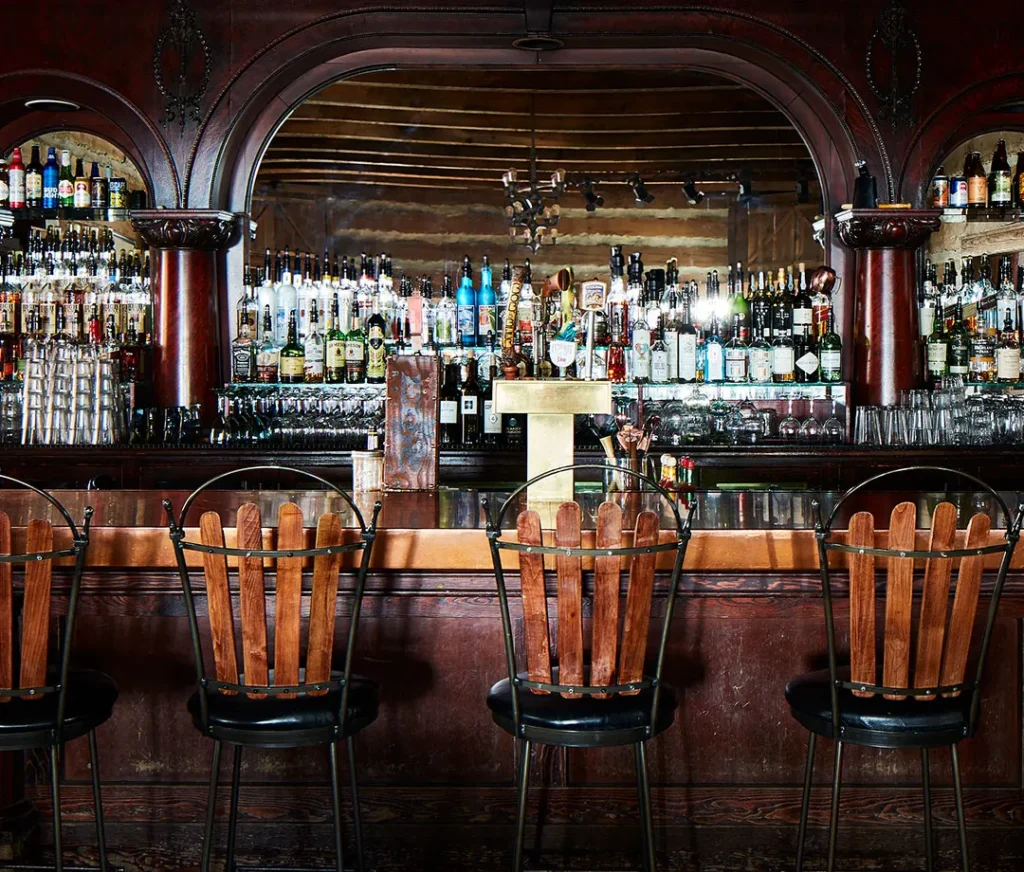 An empty, dimly-lit bar featuring an ornate wooden bar counter with four high-backed wooden stools. Behind is an extensive display of various liquor bottles in front of a large mirror and wooden shelves, creating a classic, vintage ambiance.