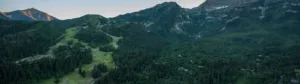 Aerial view of a vast green valley surrounded by towering, forested mountains. Sparse clusters of houses are nestled among the trees, and a winding road cuts through the landscape. The sun casts light on the mountain peaks in the background.