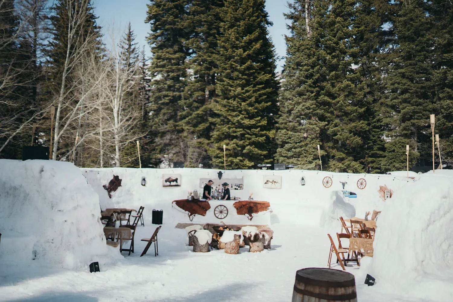 An outdoor setup in a snowy forest features rustic wooden tables and chairs arranged on snow. The area is decorated with Western-themed items like wagon wheels and animal hide rugs. Tall evergreen trees surround the space, and a clear sky is visible above.