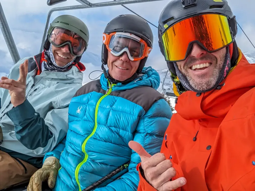 Three friends dressed in winter gear and ski goggles take a selfie on a ski lift. The person on the left is wearing a light blue jacket, the person in the middle is wearing a bright blue jacket, and the person on the right is wearing an orange jacket and reflective goggles. All are smiling and appear to be having fun. Snowy mountains can be seen in the background.