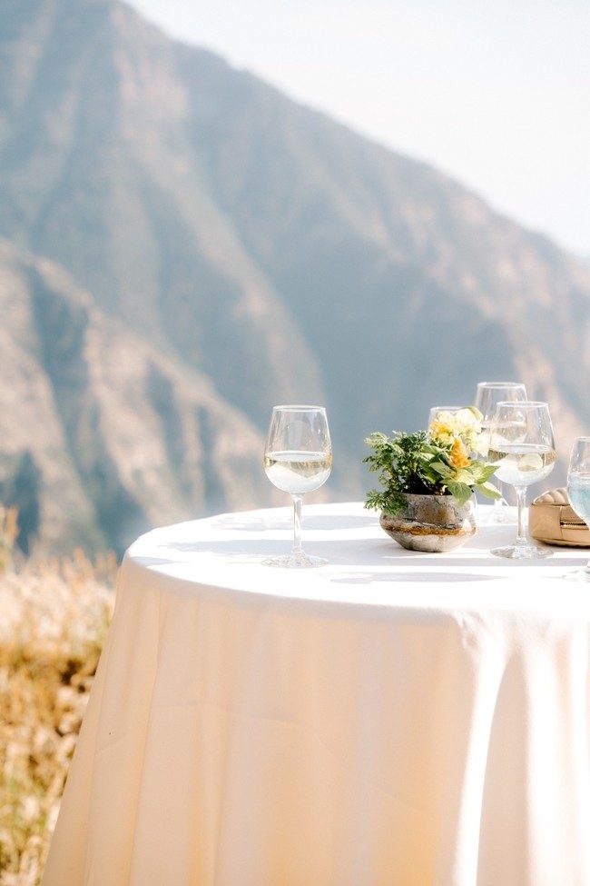 A small round table covered with a white tablecloth is set outdoors, against a backdrop of mountains. It holds two half-full wine glasses, a pair of sunglasses, and a small floral arrangement in a stone-like vase.