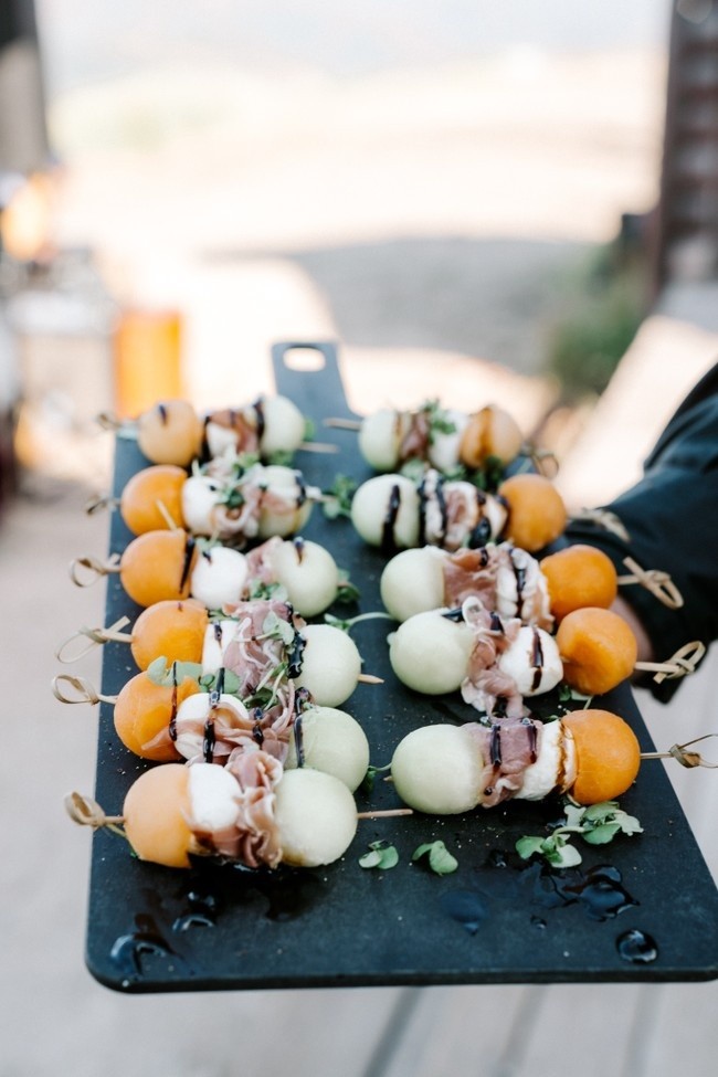 A hand holds a black tray with skewers of appetizers. The skewers contain melon balls, prosciutto, mozzarella, and fresh herbs, drizzled with balsamic glaze. The background is blurred, suggesting an outdoor setting.