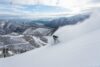 A lone snowboarder glides down a snow-covered mountain, creating a spray of powder. In the background, snow-capped mountains, valleys, and a distant lake under a partially cloudy sky offer a breathtaking view.