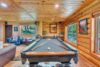 A cozy log cabin room with a pool table at the center, surrounded by comfortable leather chairs and couches. Large windows allow natural light to enter, showcasing lush greenery outside. The walls are adorned with wooden paneling and framed art.