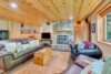 A cozy living room with wooden ceiling and walls. There are two large leather sofas, a wooden coffee table, and a leather armchair. A stone fireplace is against one wall, a TV above it. A staircase with wooden steps is in the corner, and a window overlooks outside.