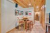 A cozy room with a wooden ceiling, tiled floor, and a round wooden table with two stools and a cushioned chair. The walls are adorned with colorful framed artwork. In the background, a hallway leads to another space with a rug and wooden furniture.