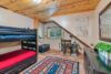 A cozy bedroom with a wooden ceiling and walls, featuring a black bunk bed. A vibrant patterned rug covers the floor. The room includes a small desk under a window, a mirror on a dresser, and a macrame wall hanging. A lamp and small decorative items are on the desk.