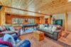 A cozy log cabin living room featuring leather sofas with cushions, a wooden chest as a coffee table, and a pool table in the background. The room has a wooden ceiling, large windows with a forest view, and various decorative items and rugs.