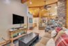 A cozy living room with a high wooden ceiling, stone accent wall, and large windows. The room features a couch, coffee table, flat-screen TV, rustic furniture, and a ceiling fan. The space opens into a dining area and kitchen with wooden cabinetry and modern appliances.