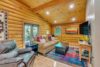 A cozy living room in a log cabin with wooden walls and ceiling. It features a beige sofa with plaid cushions, a large leather recliner, a black coffee table, a colorful rug, a flat-screen TV, and a wall painting depicting a forest scene.