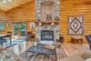 A cozy log cabin living room features a stone fireplace with rustic wooden accents. The room has wooden walls and ceiling, a geometric-patterned rug, and a large artwork on the wall. Large windows and glass doors allow natural light to brighten the space.