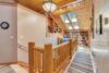 A cozy wooden hallway featuring a rustic railing, a large chandelier, and skylights providing natural light. Shelves filled with books and decor are visible at the far end, with striped rugs running along the light wooden floor. A clock hangs on the left wall.