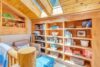 A cozy reading nook with wooden shelves filled with books and decor, bathed in natural light from skylights. There is a leather armchair and a blue ottoman. A blanket is draped over the armrest, and plants add a touch of green to the warm space.