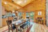 A warm, rustic dining room with wooden log walls and high ceilings. A long wooden dining table with chairs and benches is in the center. The room is illuminated by natural light from large glass doors leading to a balcony with forest views. The kitchen is in the background.
