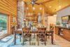A cozy dining and living area featuring a rustic wooden table with six chairs. A stone fireplace and plush seating dominate the background. The space boasts wooden walls and ceilings, ceiling fans, and warm lighting. Large windows let in natural light and offer outdoor views.