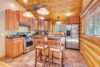 Rustic kitchen with wooden cabinetry and log walls, stainless steel appliances, and a tiled floor. An island with two wooden barstools is in the center, and there are large windows bringing in natural light. Pendant lights hang over the island.