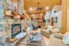 A cozy wooden cabin living room with a stone fireplace on the left, comfortable beige sofas, a coffee table with decor, and large windows providing natural light. The room features wooden walls and ceiling with a fan, creating a warm and inviting atmosphere.