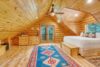 A cozy, rustic bedroom with wooden log walls and ceiling. The room features a large bed with white bedding, wooden dressers, a ceiling fan, and a vibrant blue and red patterned rug. Double glass doors open to a balcony overlooking a forested area.